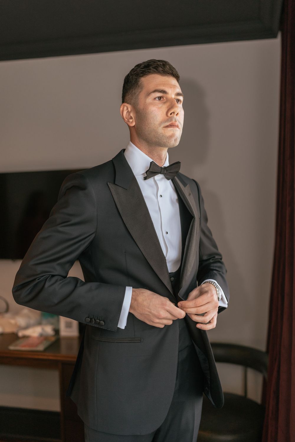 A young man in a black tuxedo with bow tie buttons his jacket while standing in an elegant room.