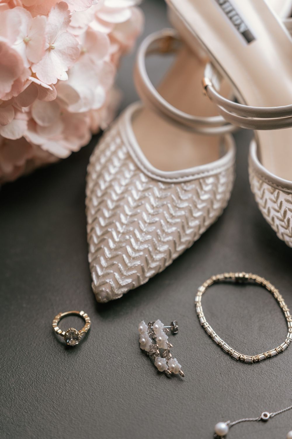 Elegant bridal accessories flat lay with ivory heels, crystal earrings, gold ring, bracelet, and pink hydrangea on dark background.