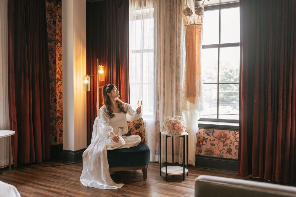 Bride in white robe sitting in elegant hotel suite chair, holding phone near large window with wedding dress hanging nearby.