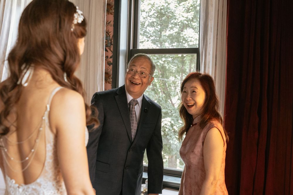 Bride in white gown shares a joyful first look moment with smiling parents near a window.