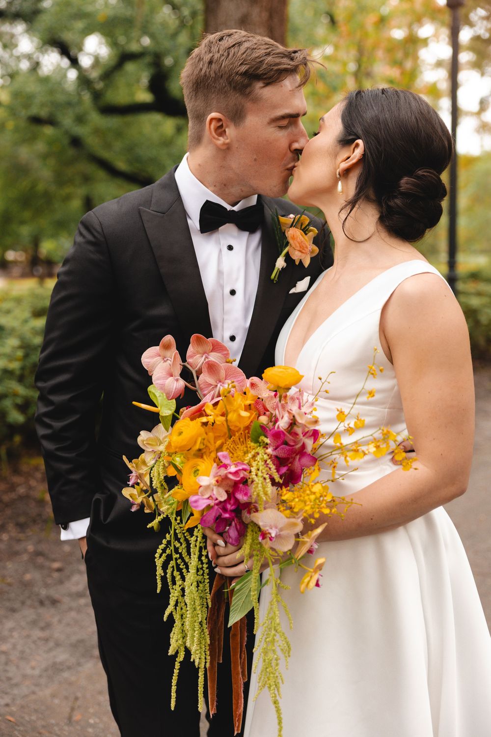 Bride in a white gown and groom in a black tuxedo share a kiss outdoors while holding a colorful wedding bouquet of orange and pink flowers.