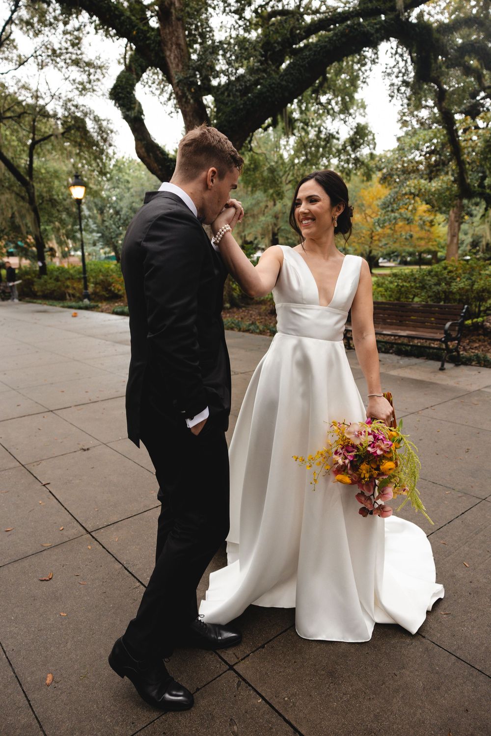 Groom in a black suit kisses the hand of his smiling bride in a white gown holding a colorful bouquet in a tree-lined park.
