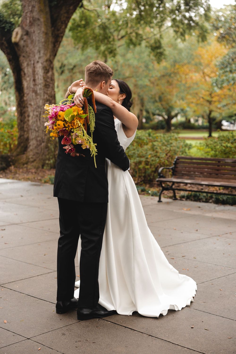 A bride in a white gown and a groom in a black suit embrace in a park, holding a colorful autumn bouquet near a bench and trees.
