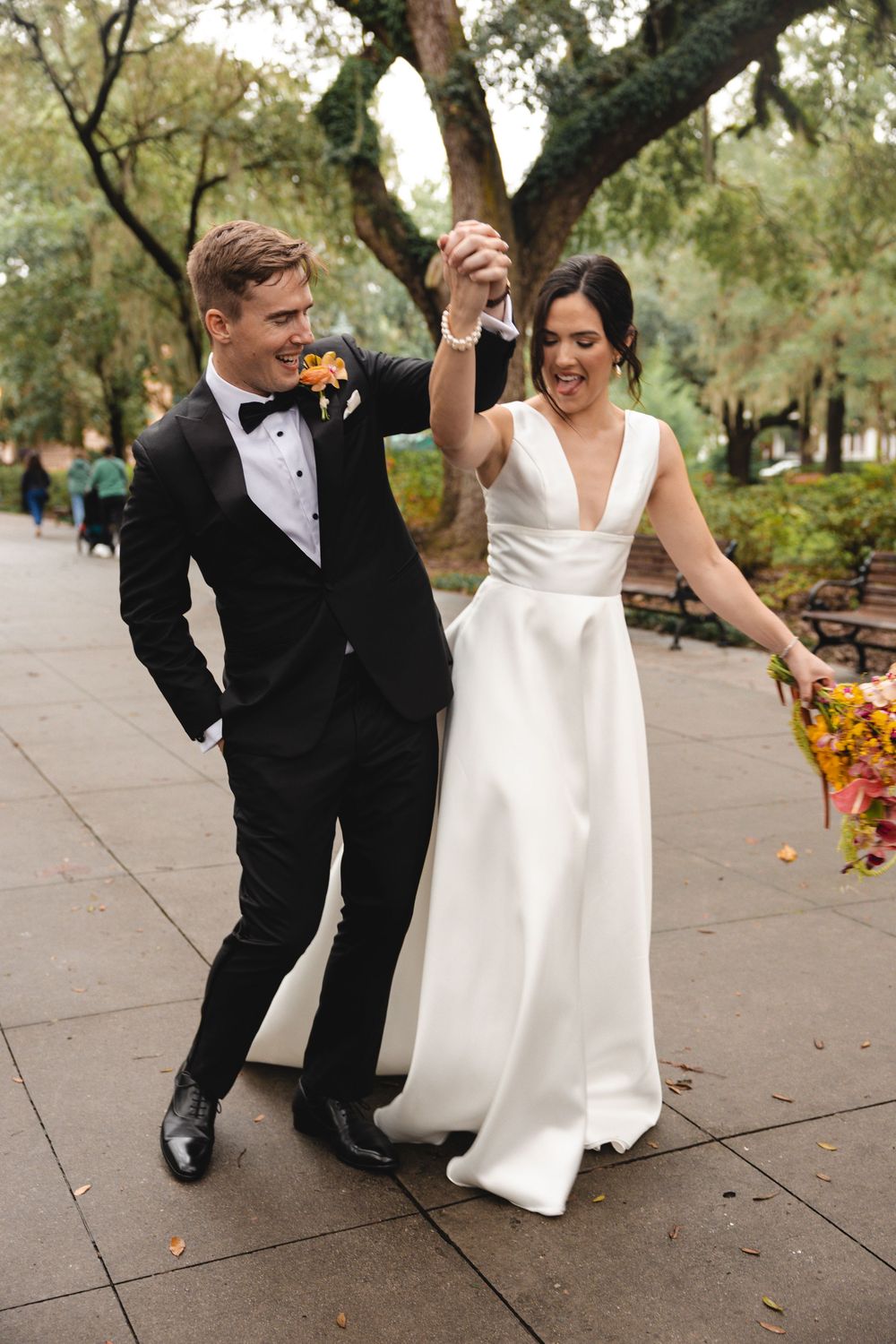 Smiling bride in a white V-neck gown and groom in a black tuxedo hold hands raised, walking joyfully beneath oak trees after their wedding.