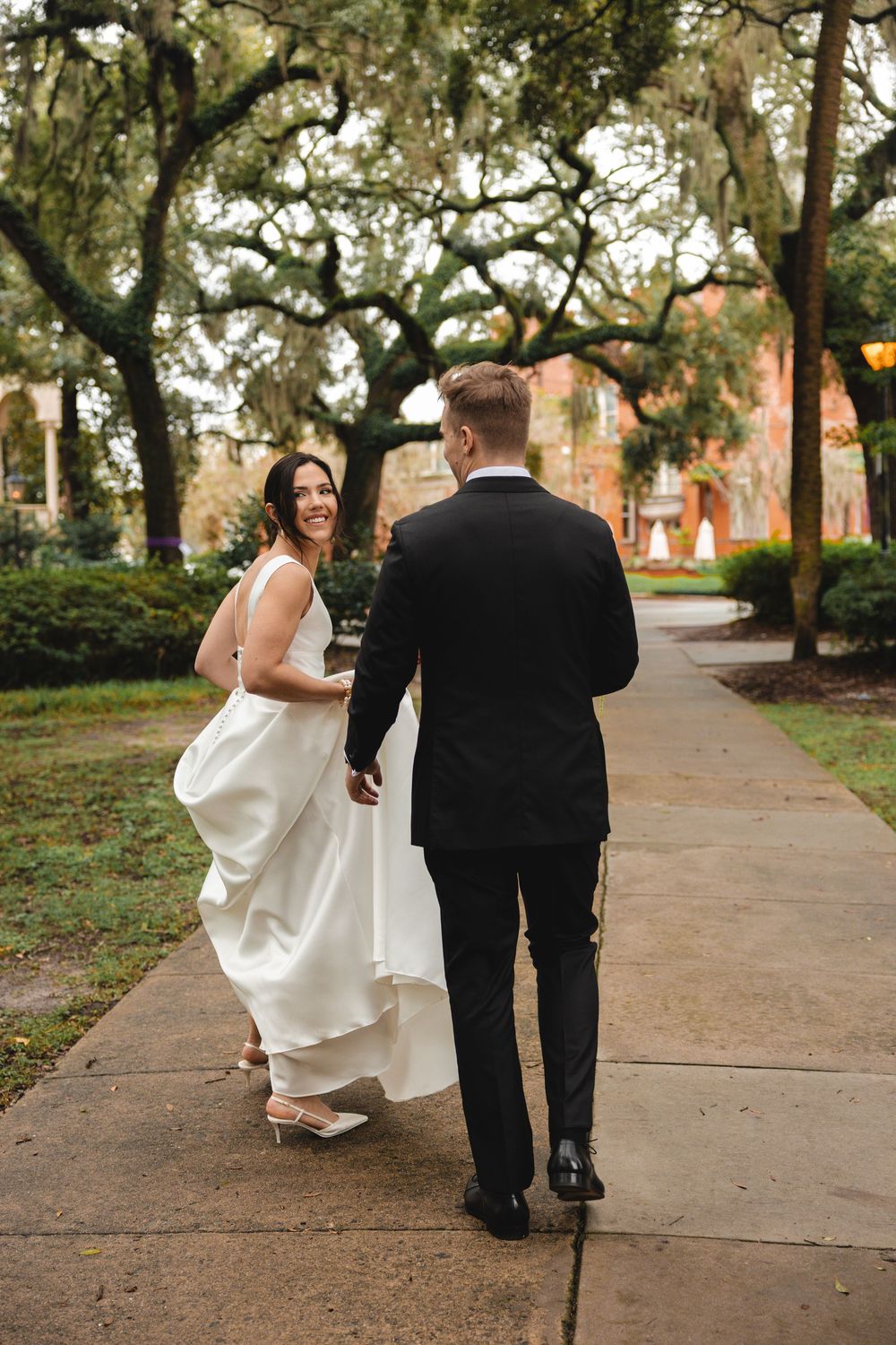 Bride in a white gown smiles back over her shoulder while walking hand in hand with a groom in a black suit under oak trees.