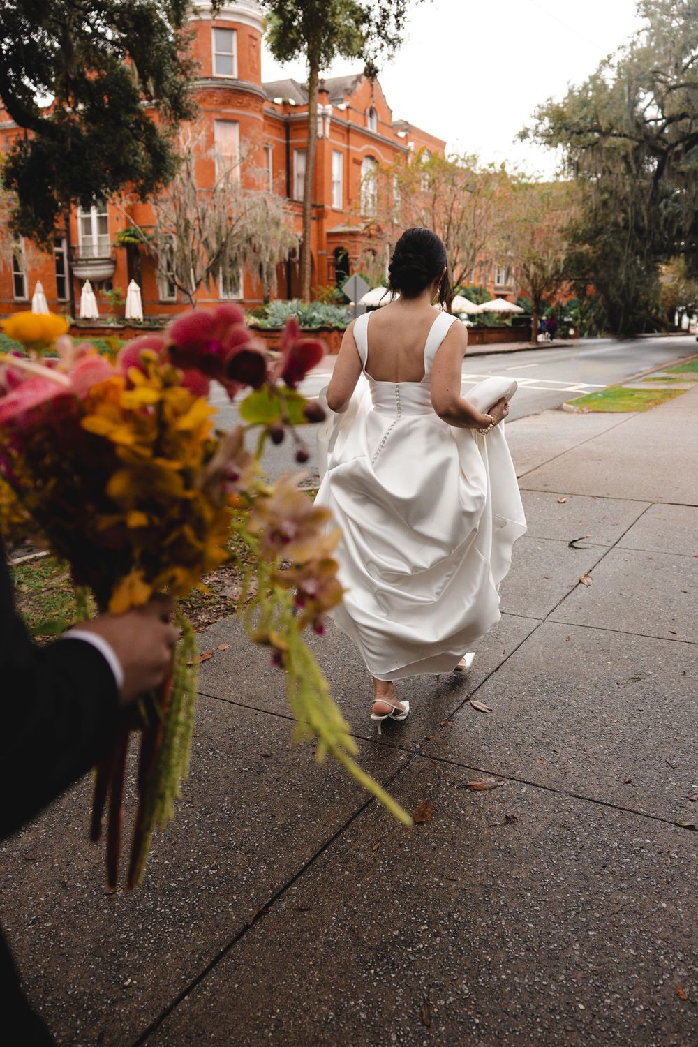 Bride in a flowing white gown walks away down a tree-lined sidewalk toward a historic red brick building, holding a colorful bouquet.
