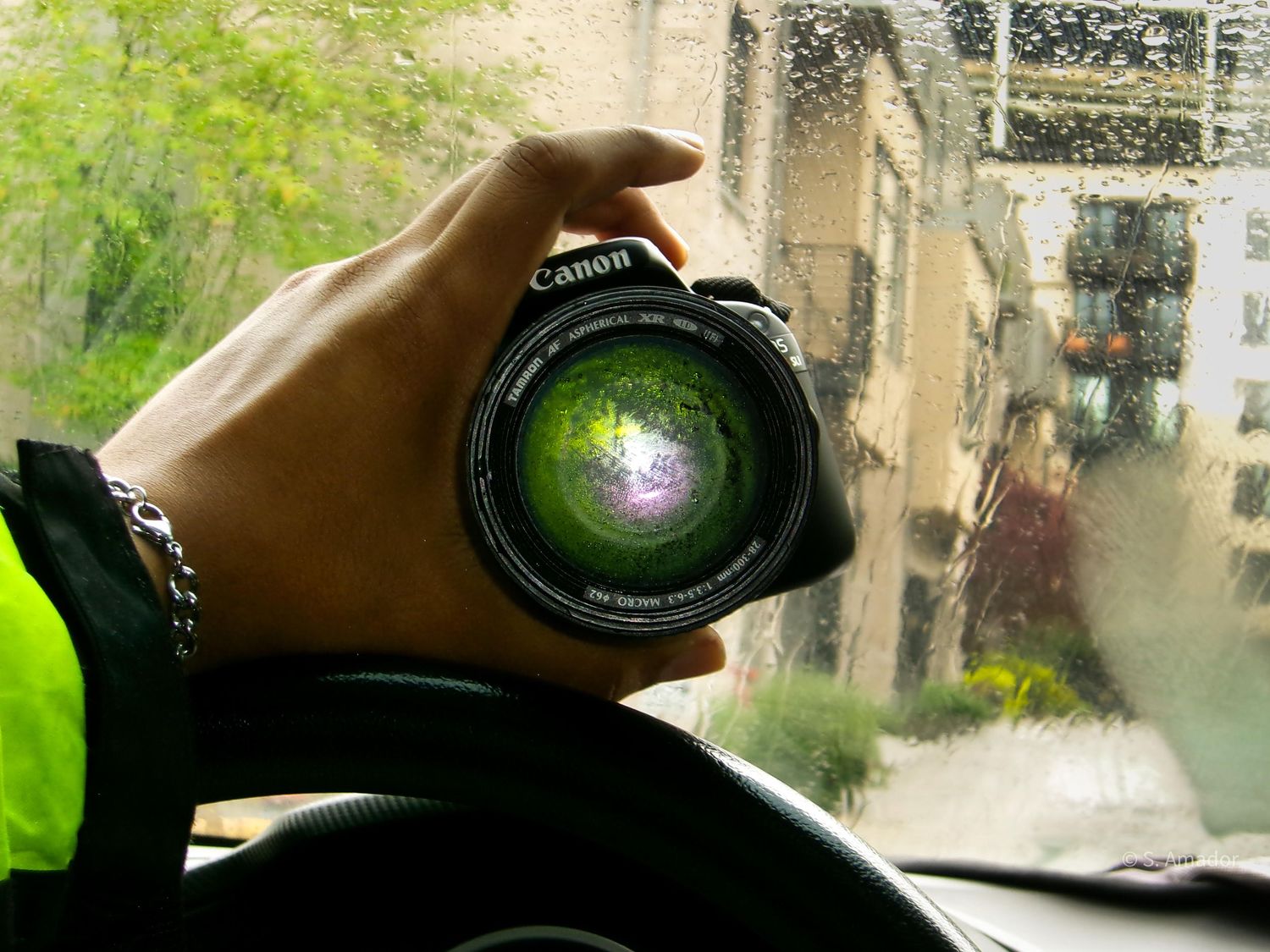 Hand holding a Canon camera lens inside a car with rain-covered window in background.