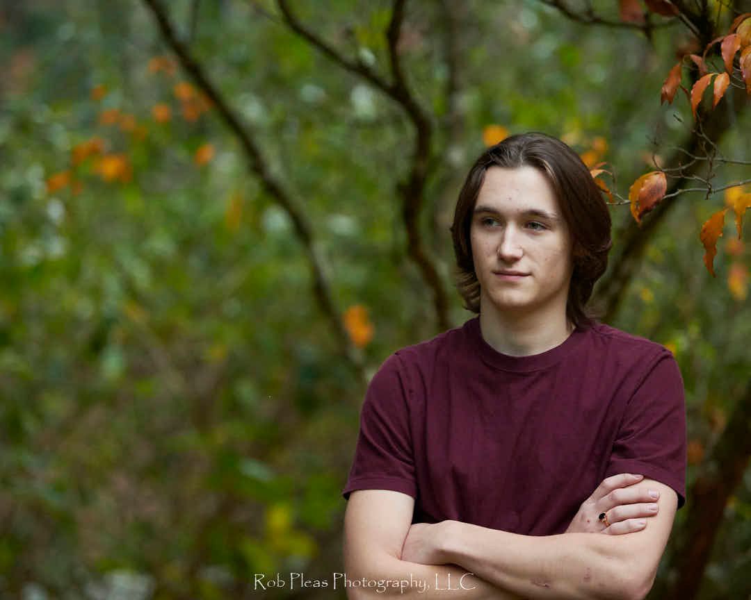 High school senior male with arms crossed, standing in front of tree with fall-colored leaves.