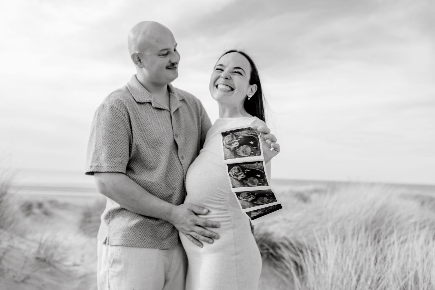Black and white photo of a smiling pregnant couple on a beach holding ultrasound scans.