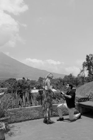 Black and white photo of a man proposing on one knee to a woman with a volcano backdrop.