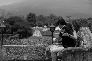 Black and white photo of a couple embracing on a stone wall with mountain landscape in background.