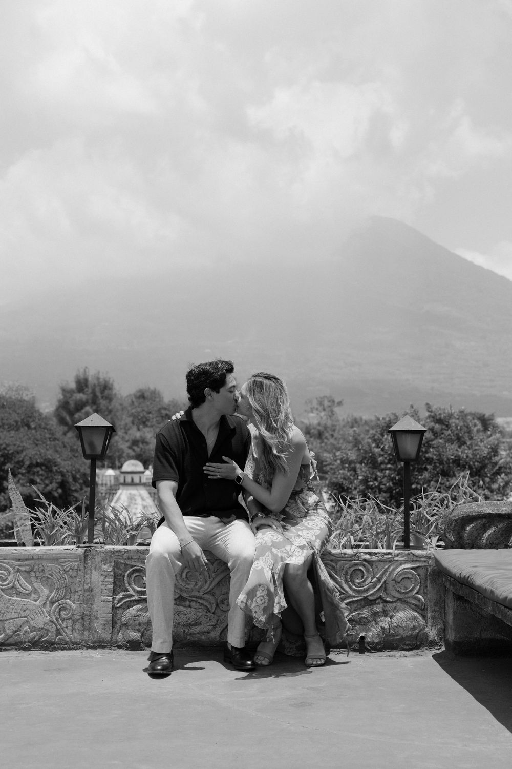 Black and white photo of a romantic couple kissing on a stone wall with a volcano and cloudy sky in the background.