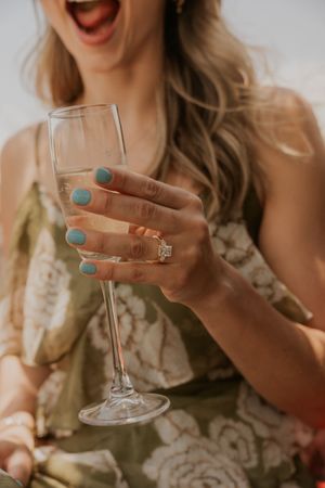 Woman in floral dress holding champagne flute with diamond engagement ring and teal nail polish, mouth open in excitement.