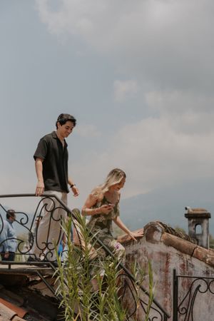 Couple exploring ancient ruins on a rooftop with ornate iron railings and mountain views under a cloudy sky.