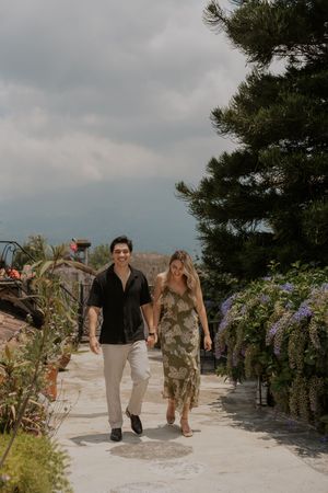 Couple holding hands walking along a garden path surrounded by lush greenery and purple flowers under a cloudy sky.