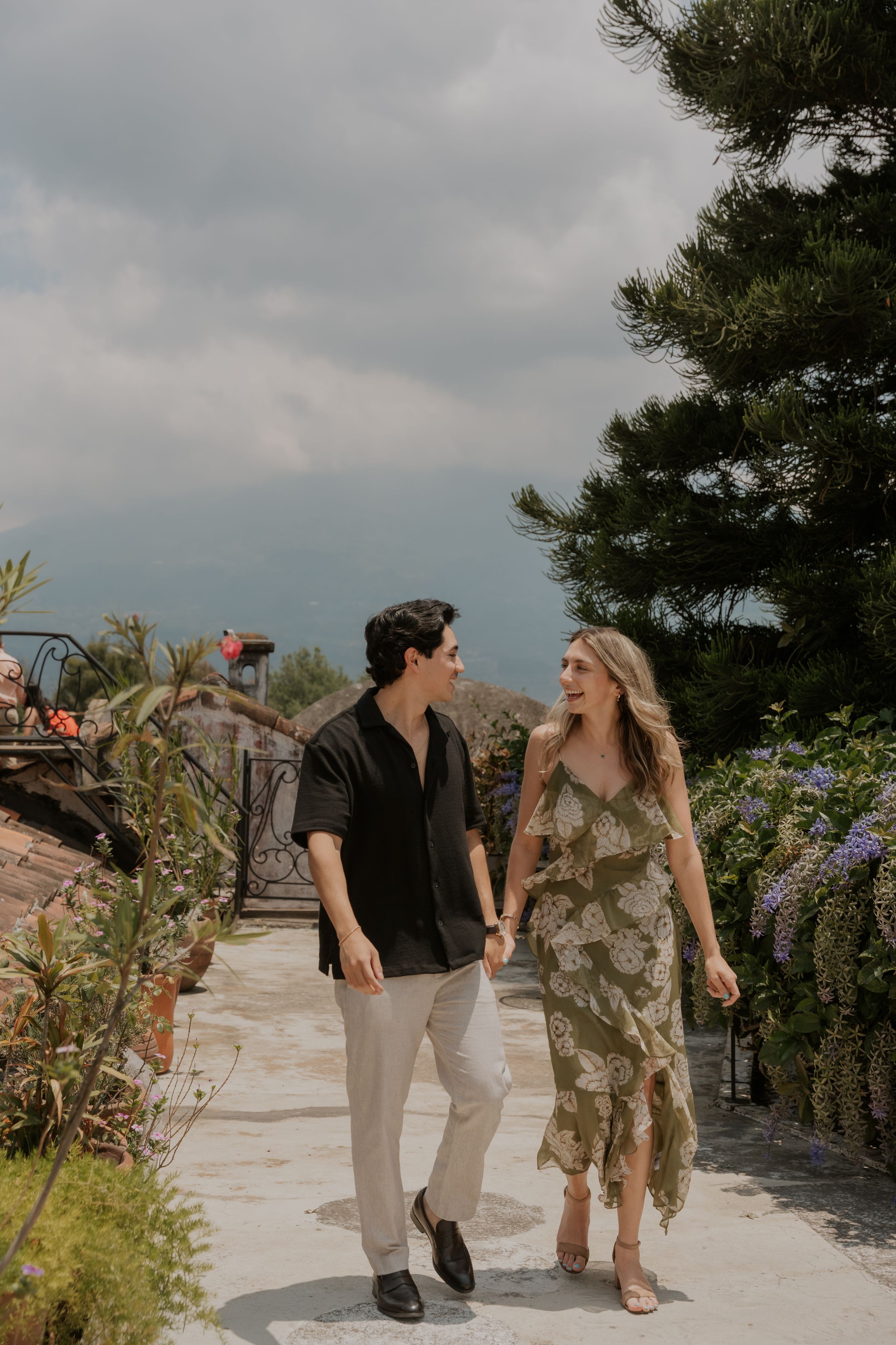 Couple holding hands and walking through a lush garden path with stormy skies in the background.