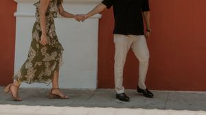 A couple holds hands while walking together, the woman wearing a floral dress and the man in white pants near a red wall.
