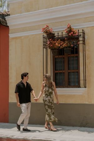 Couple holding hands walking past a colorful colonial building with flower-adorned iron window grilles.