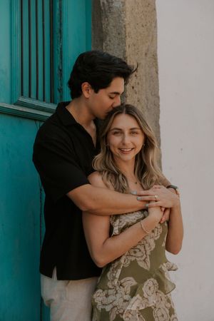Couple embracing by turquoise door, woman smiling in floral dress as man kisses her cheek.