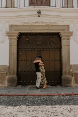 Couple embracing in front of a large ornate wooden door on a cobblestone street in a historic building.