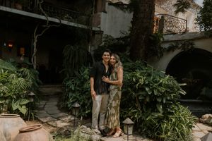 Couple embracing and smiling in a lush garden courtyard with tropical plants and stone architecture.