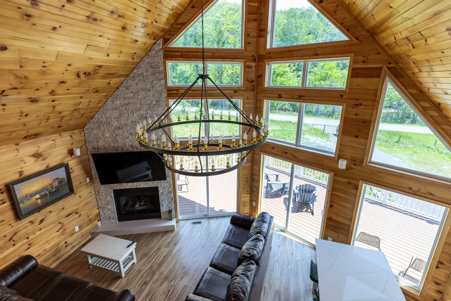 Aerial view of a rustic log cabin living room with vaulted ceilings, stone fireplace, chandelier, and large windows overlooking nature.