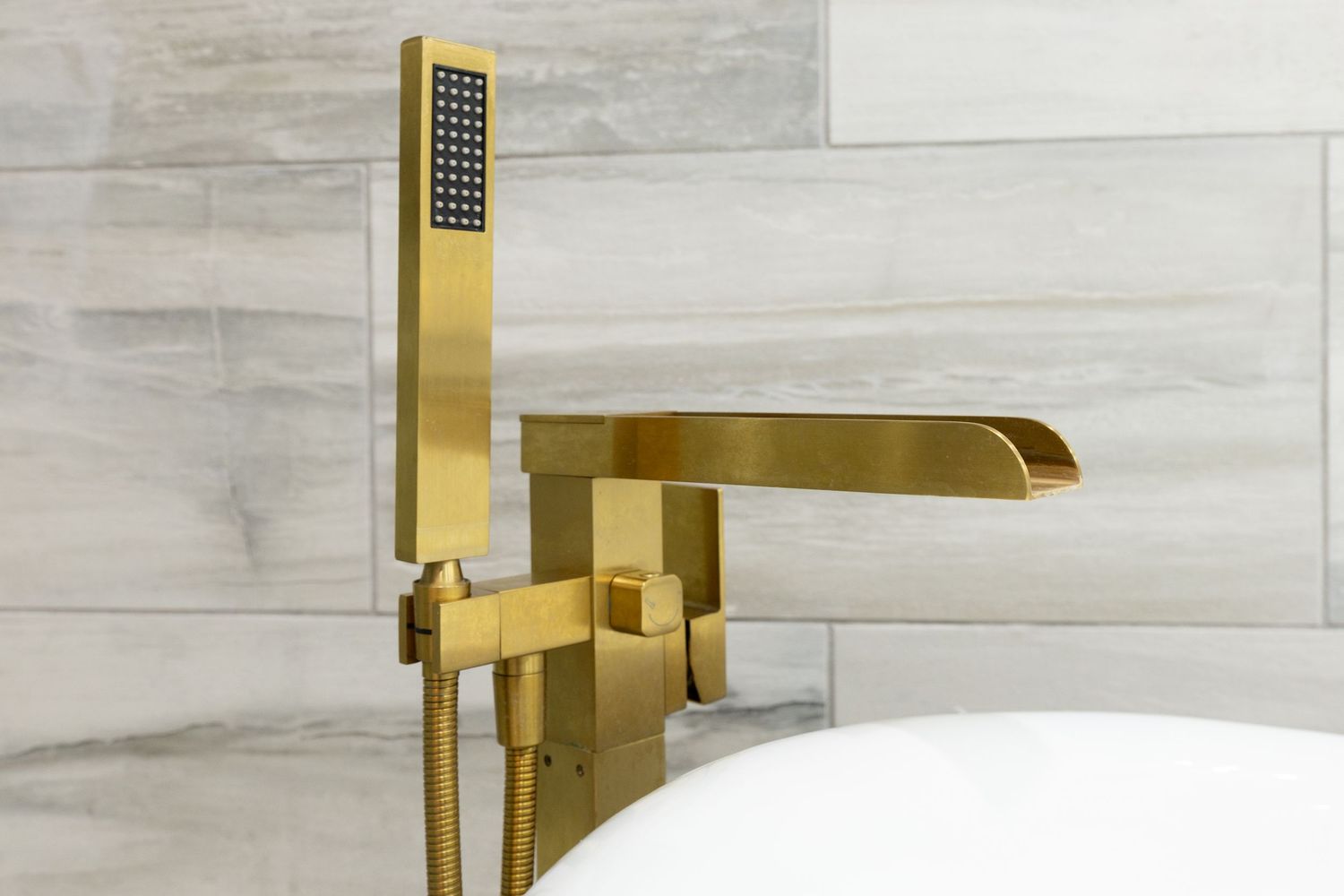 Brushed gold freestanding tub filler faucet with handheld shower head against gray tile wall.