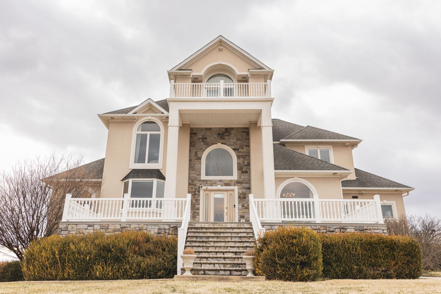 Large two-story stucco home with stone accents, arched windows, white railings, and grand staircase entrance under cloudy skies.