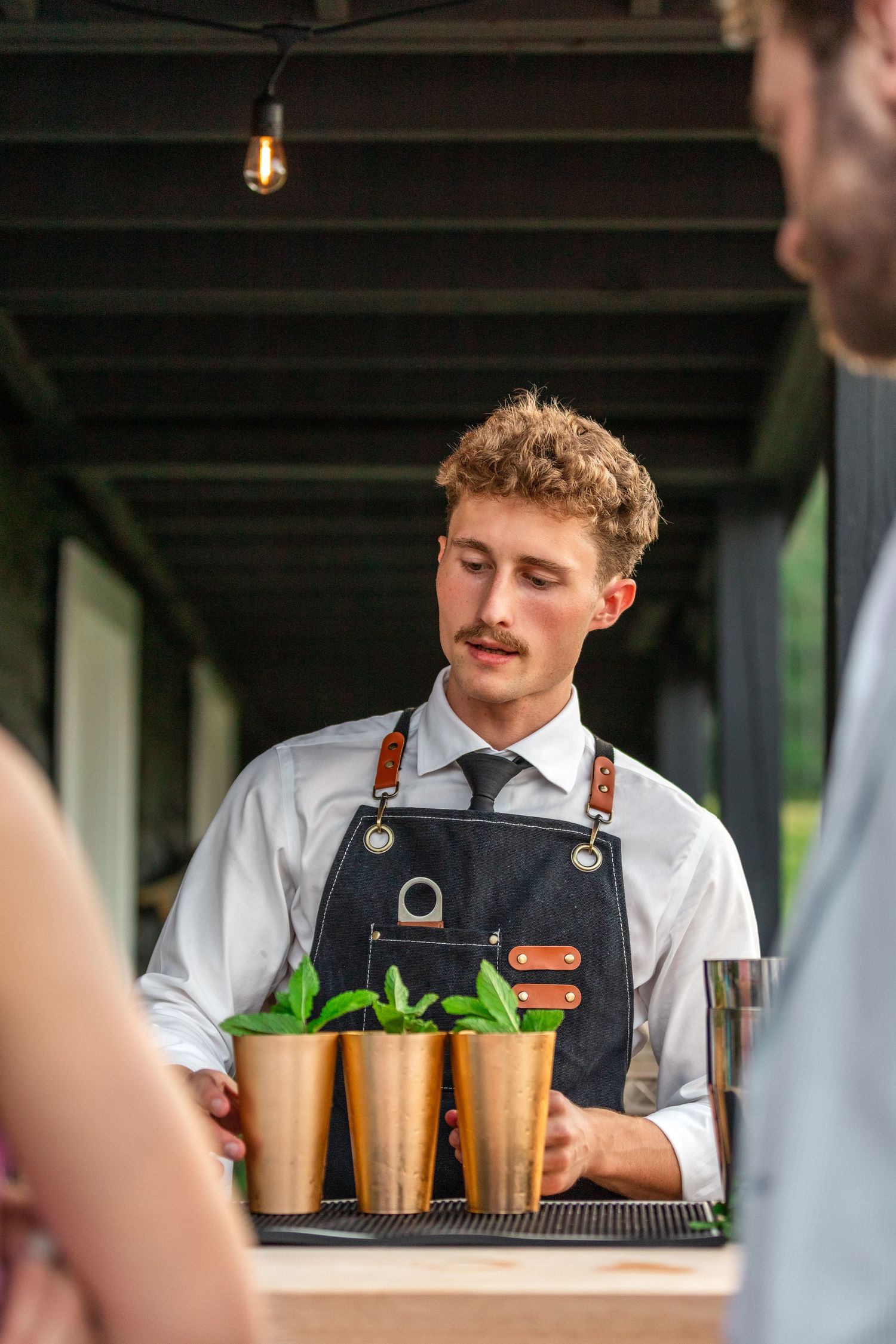 Bartender in apron prepares mint juleps in copper cups at an outdoor bar.