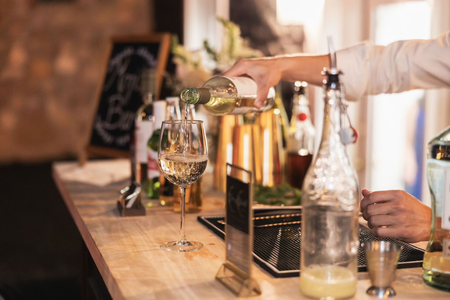 Bartender pouring white wine into glasses at a wooden bar with bottles and cocktail shaker nearby.