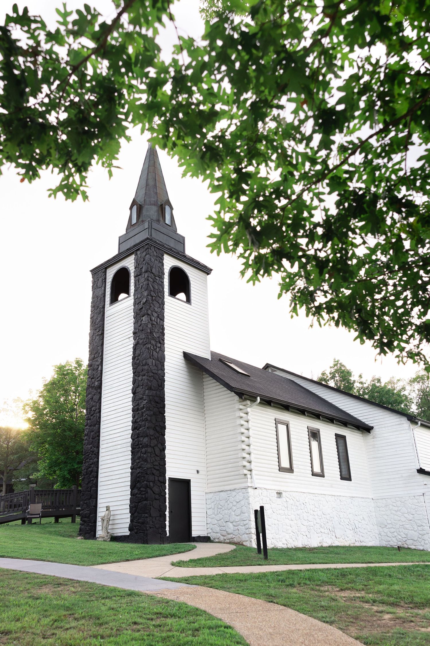 White historic chapel with dark stone bell tower and steeple, framed by green oak tree branches at sunset.