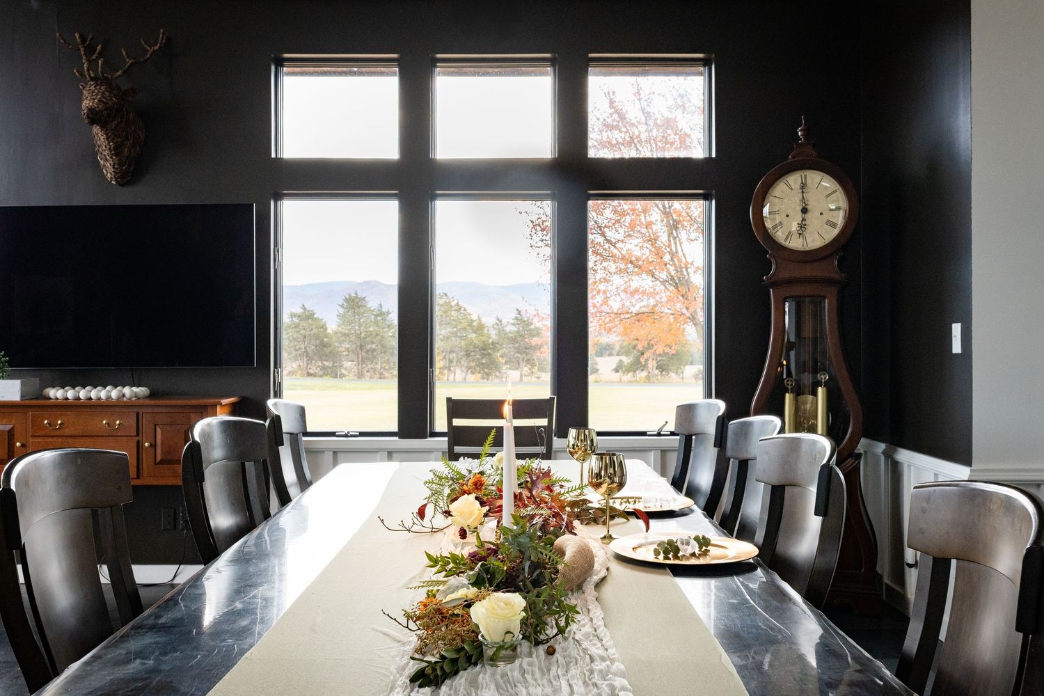 Elegant dining room with dark walls, long marble table, floral centerpiece, grandfather clock, and scenic window view.