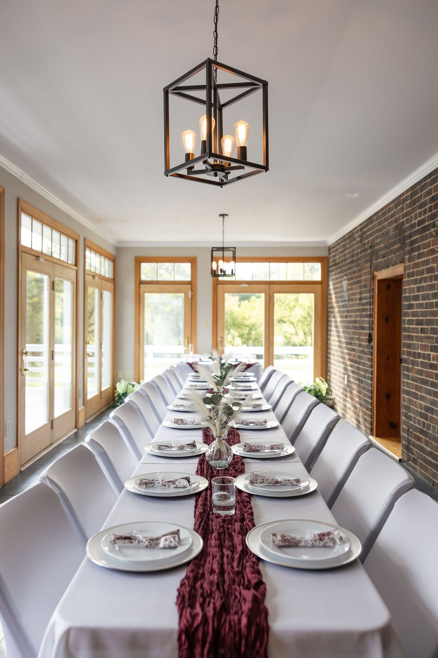 Elegant long dining table set for wedding reception with white linens, burgundy runner, and chandelier lighting in brick venue.