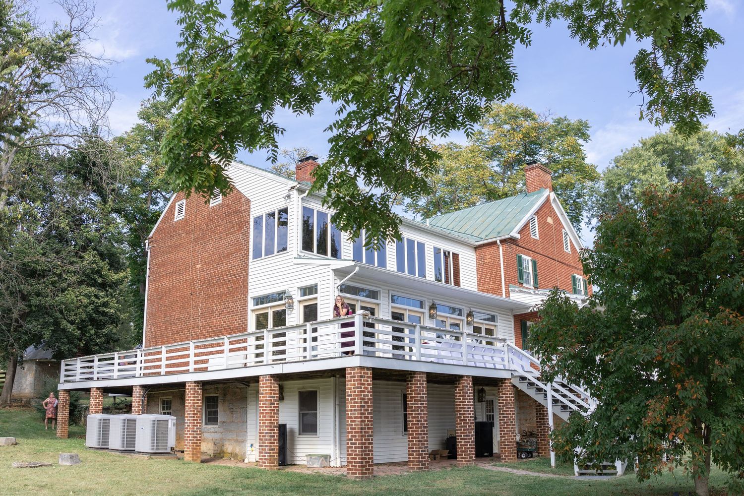 Large brick and white two-story house with green roof, wraparound deck, and mature trees.