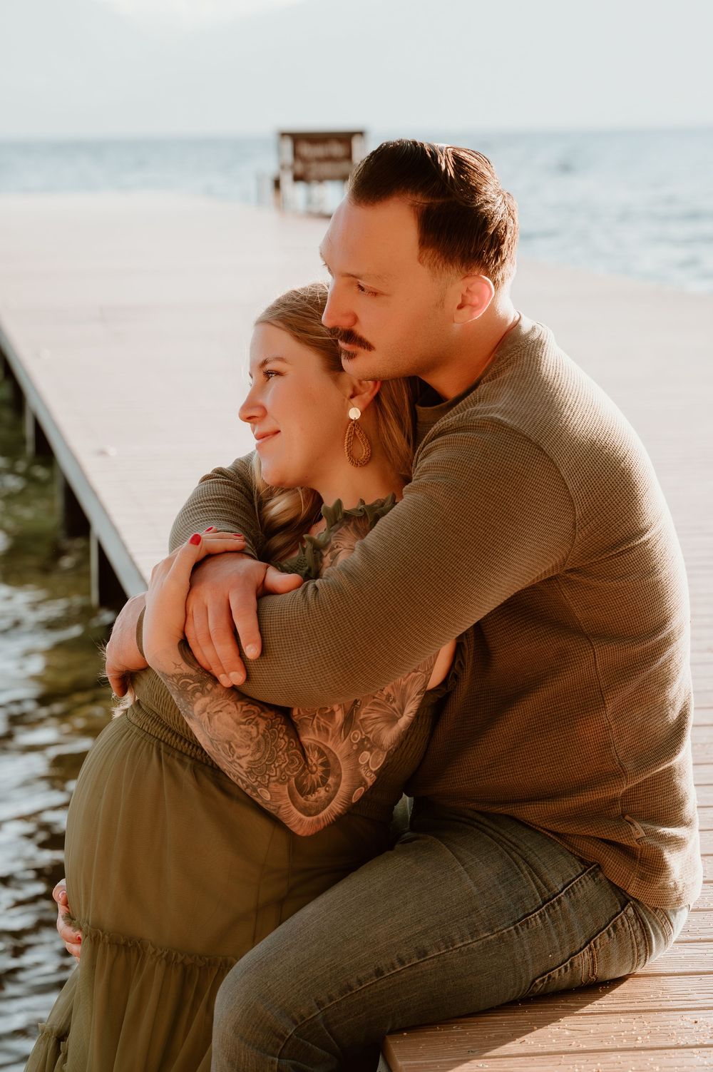 A couple sits embracing on a wooden dock by a lake, the man wrapping his arms around the tattooed woman at golden hour.