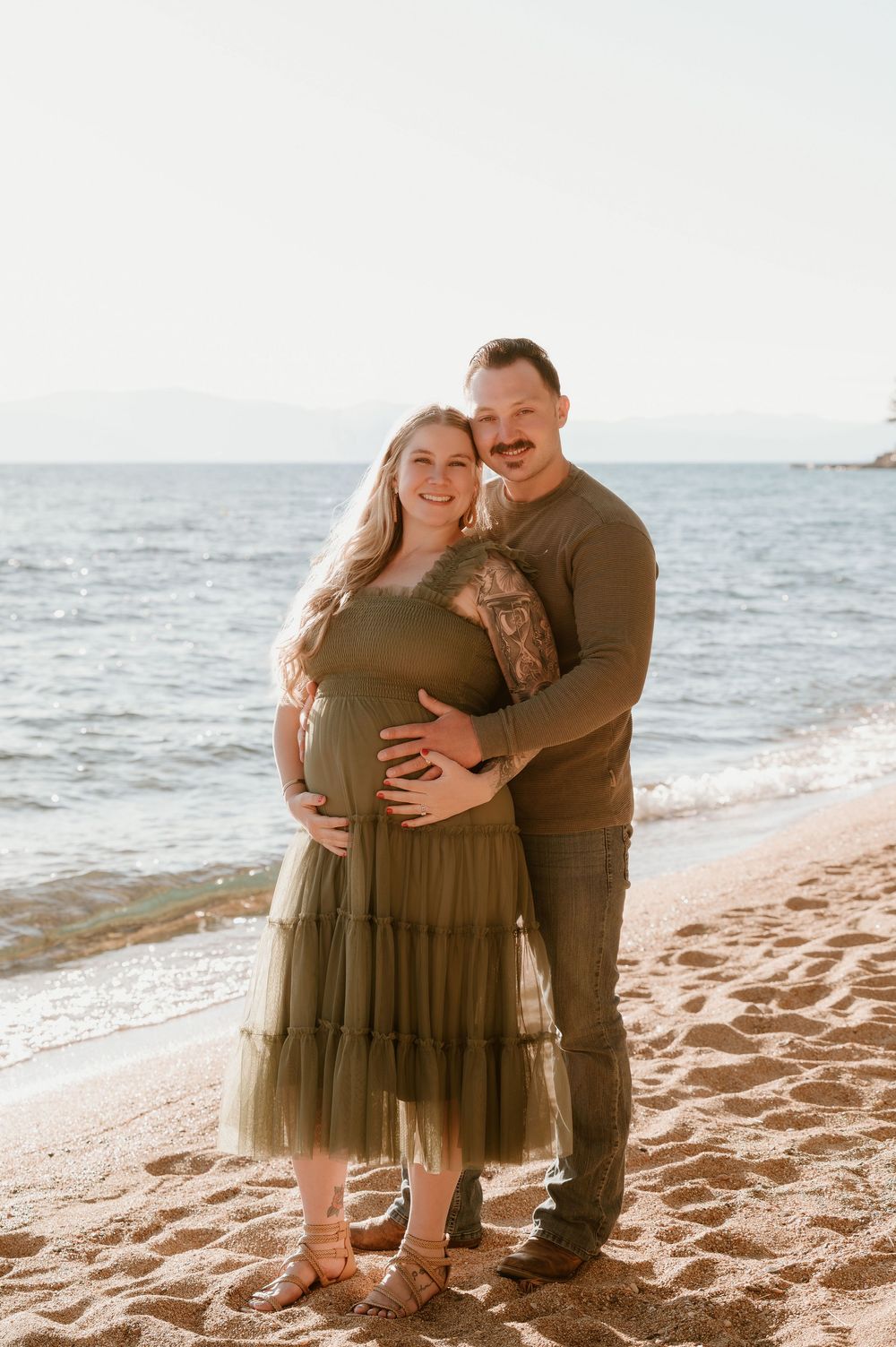 Couple in matching olive outfits posing on sandy beach, woman in green tulle dress cradling baby bump, man embracing her from behind.