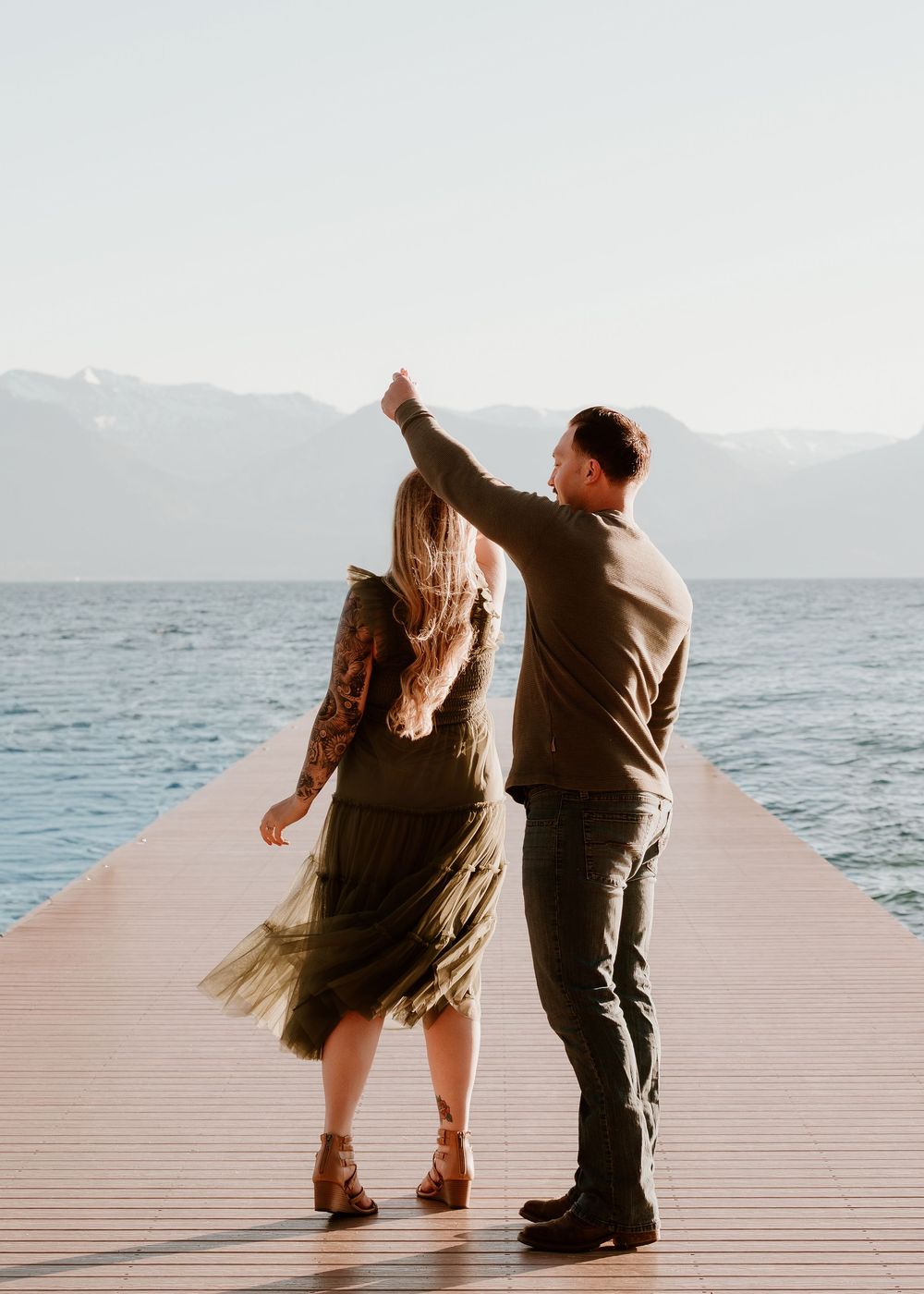 Couple dancing on a wooden pier with mountains and blue lake in the background on a sunny day.
