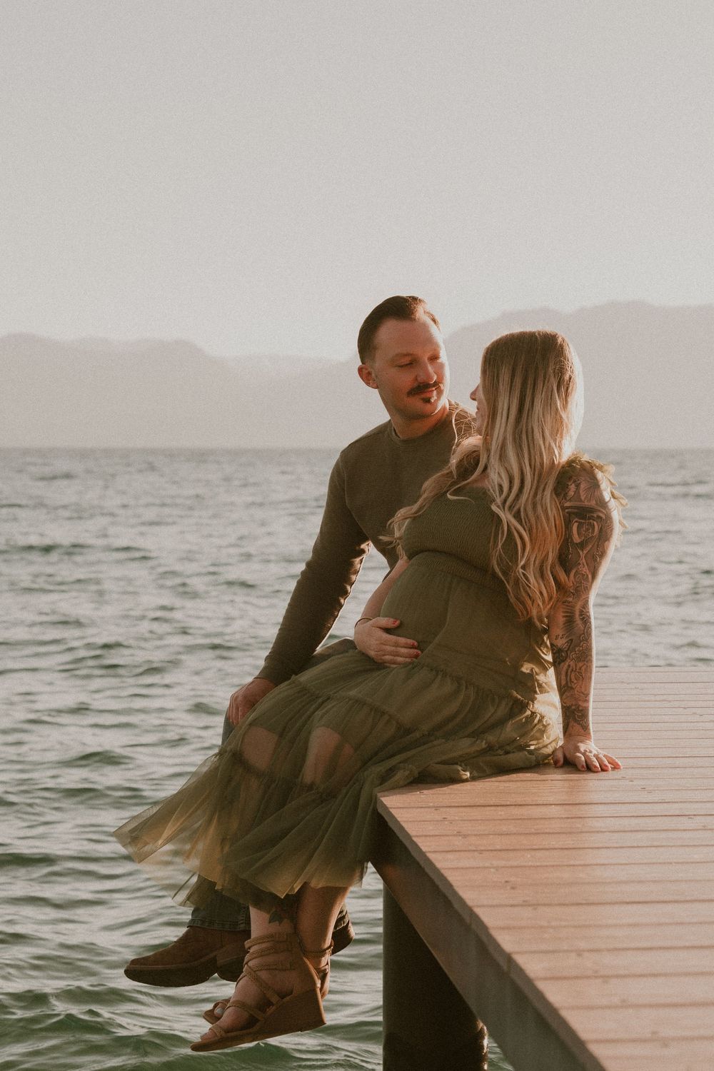 Couple in matching olive green outfits sitting on a dock by a lake, sharing a tender moment during a maternity photoshoot.