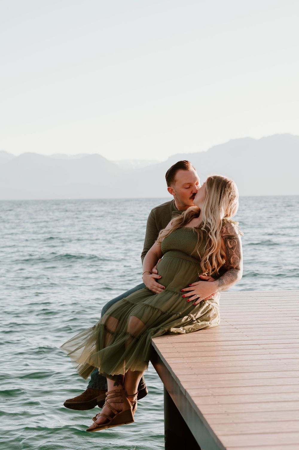 Couple sharing a kiss on a lakeside dock, woman in green maternity dress with mountains in background.
