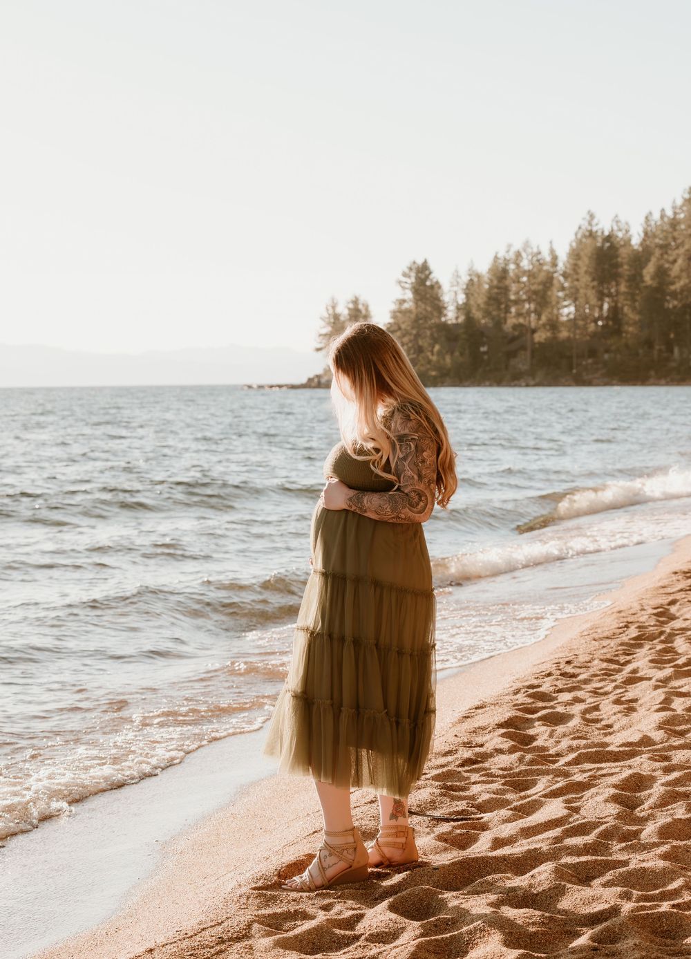 Pregnant woman in olive green tiered maxi dress standing on sandy beach shore with calm ocean waves and trees in background.