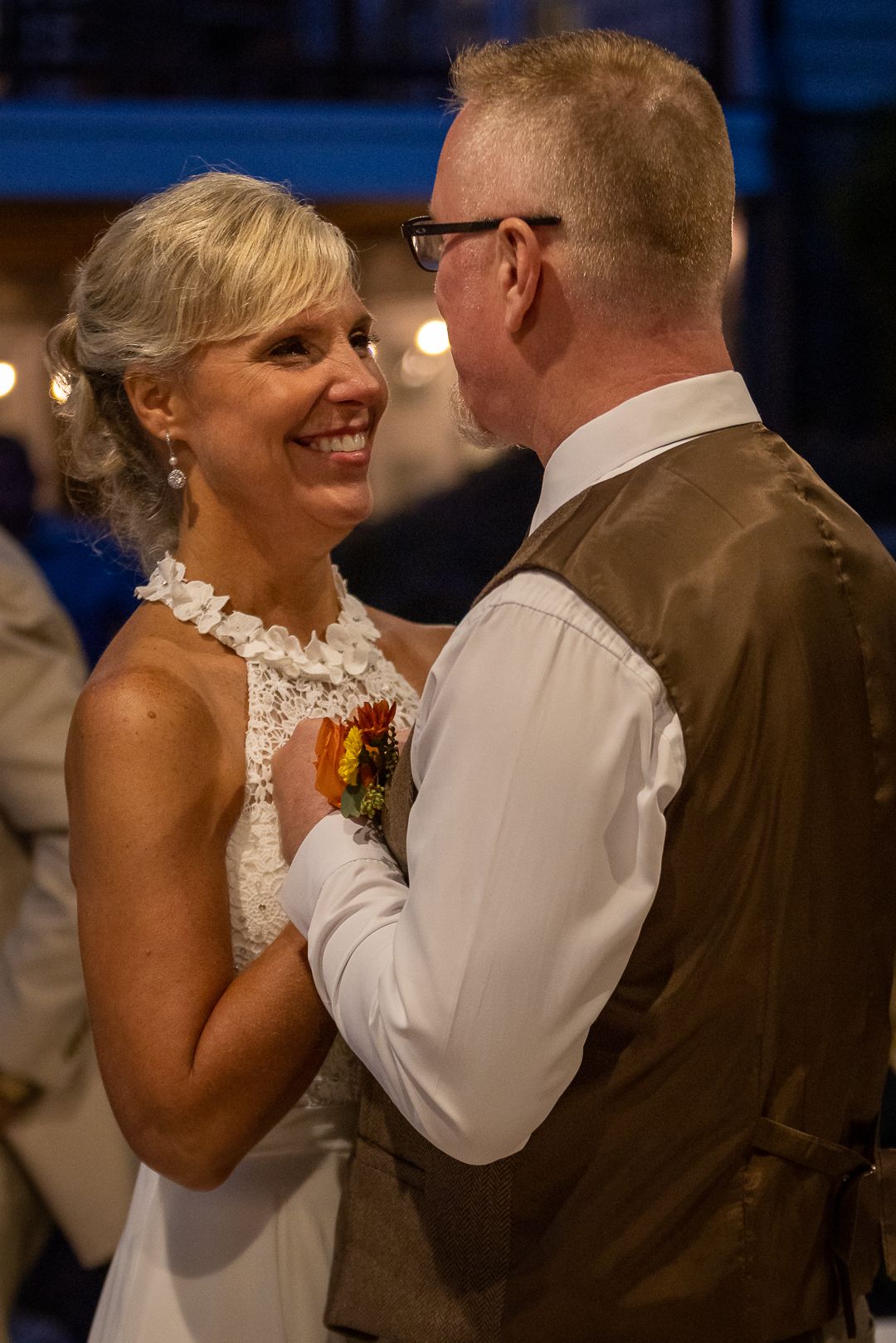 Happy newlyweds pose outdoors at fall wedding ceremony, bride in white halter gown holding orange bouquet aloft, groom in brown vest.