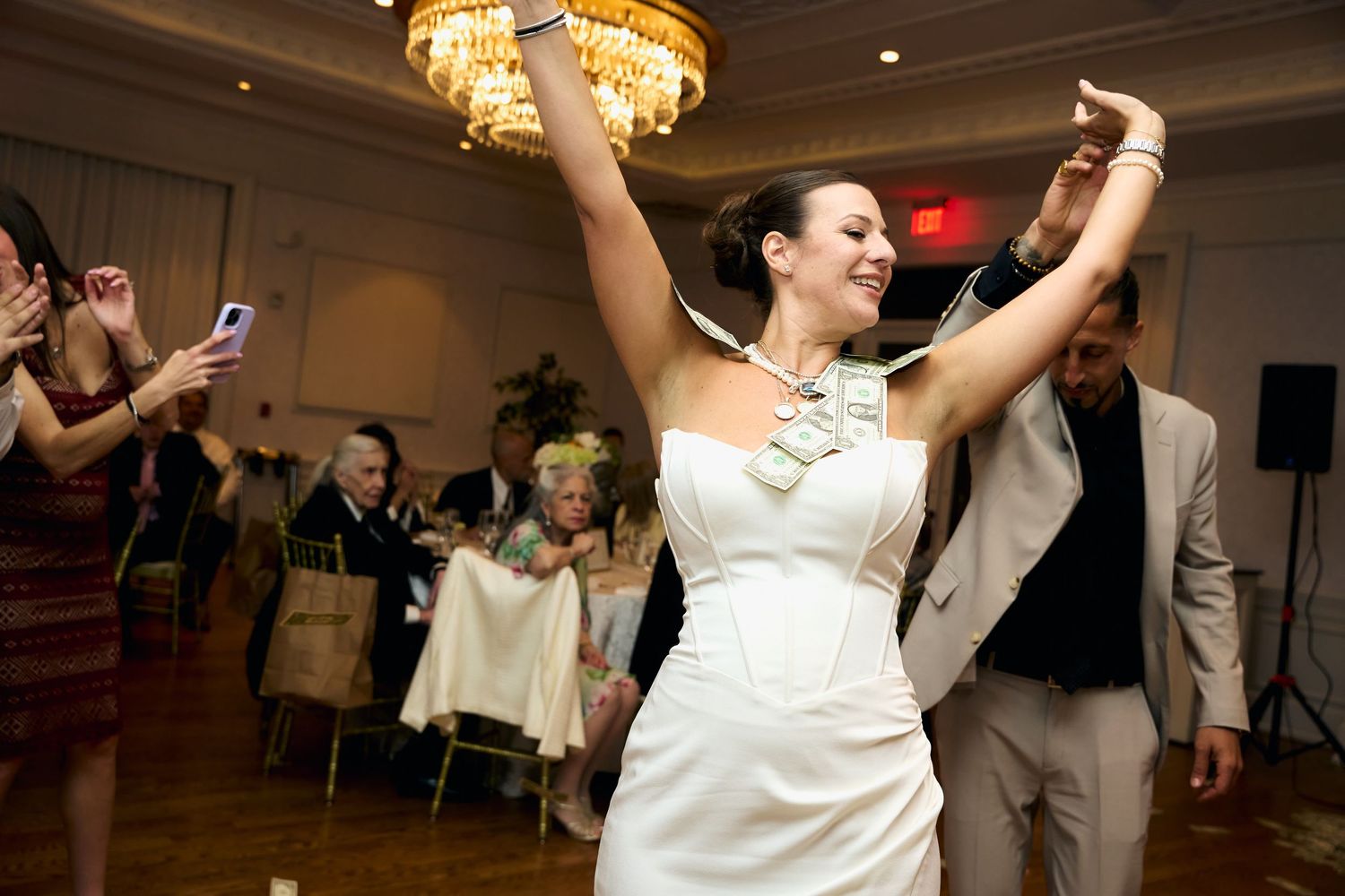 Bride in white strapless gown raises arms joyfully while dancing at elegant wedding reception ballroom.