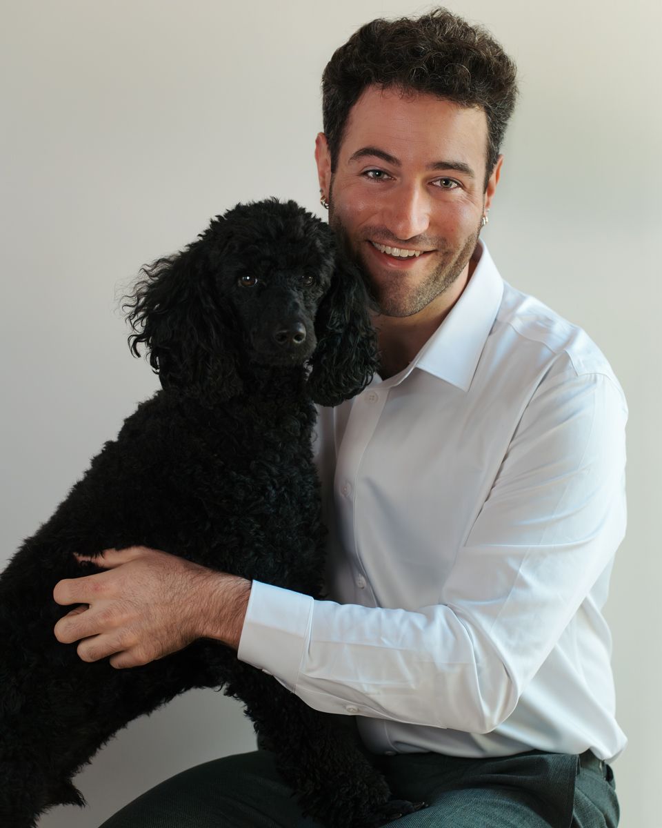 Smiling man in white shirt hugging a black poodle dog against a light gray background during an in home photoshoot in the hills Sydney