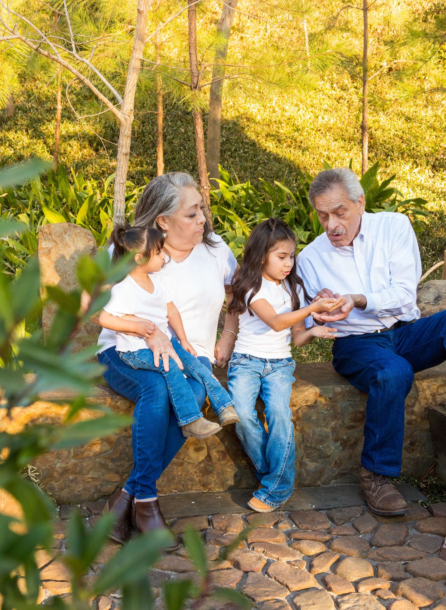 Grandparents sitting on stone bench with two young girls in a sunny park, all wearing white shirts and jeans.