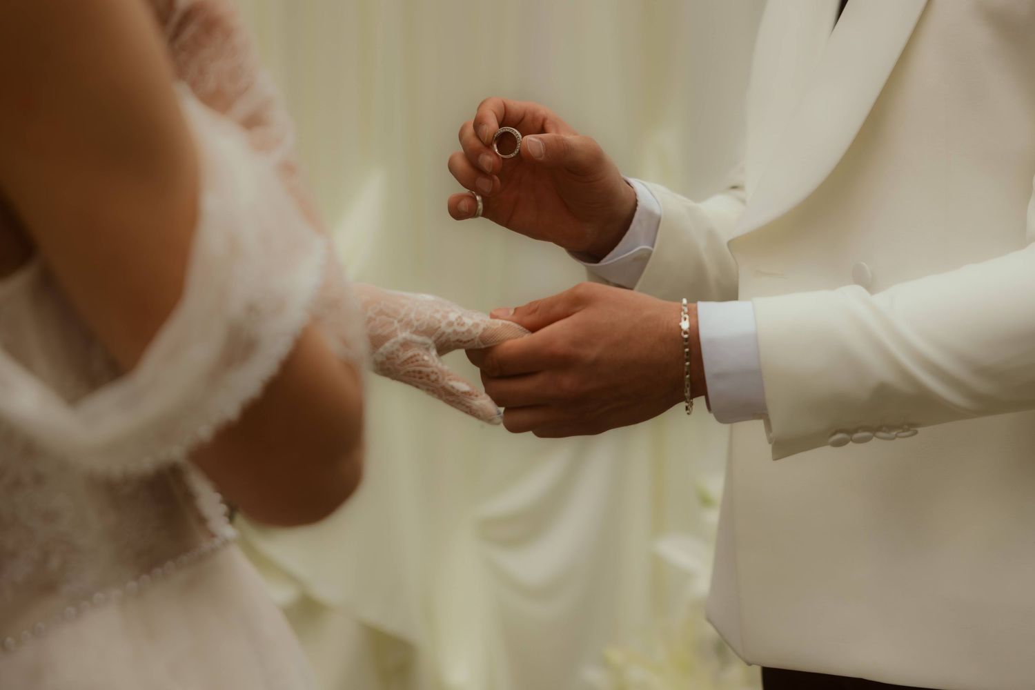 Groom in white suit placing wedding ring on bride's finger during ceremony.