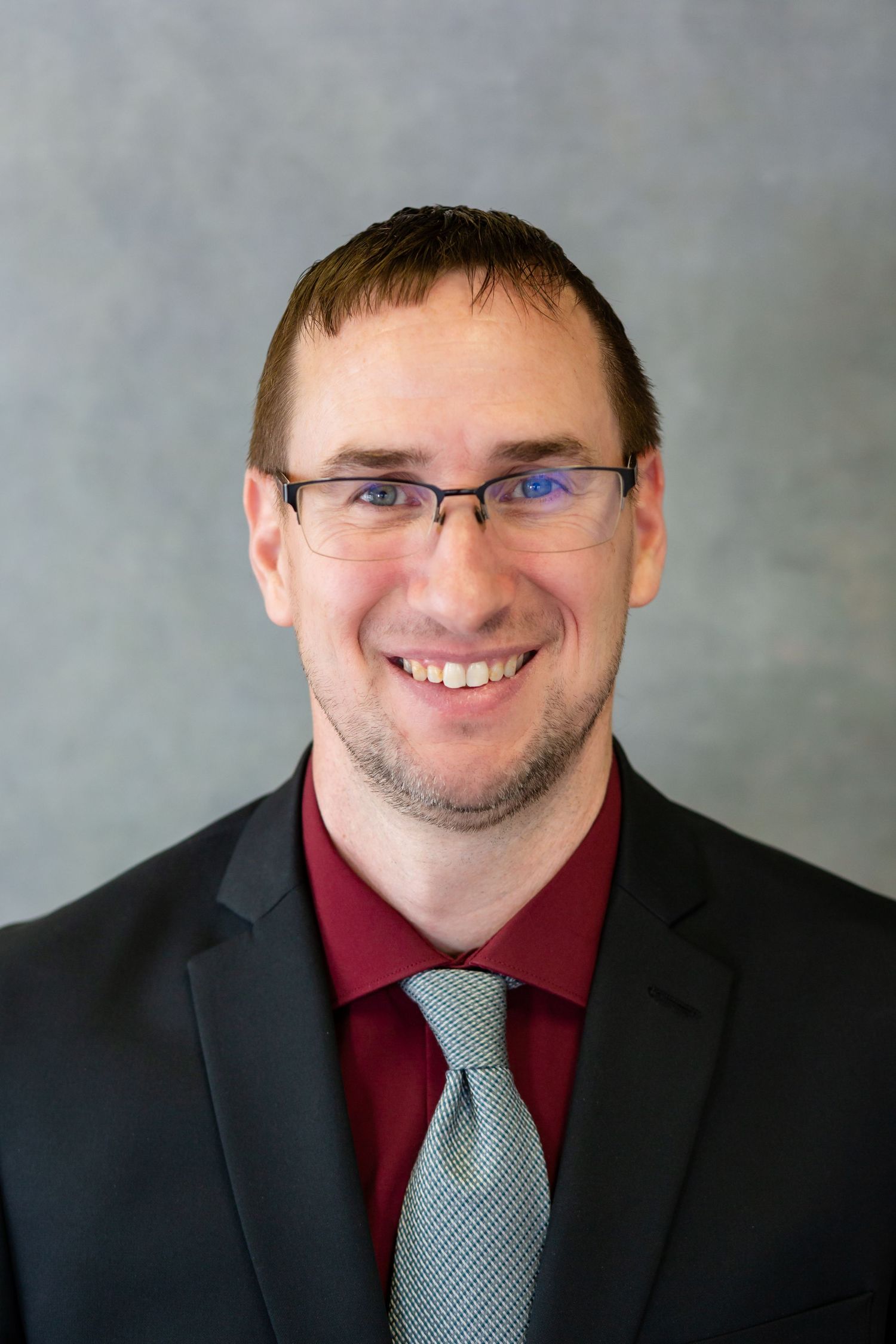 Professional headshot of a smiling man wearing glasses, burgundy shirt, gray tie, and black blazer against a gray background.