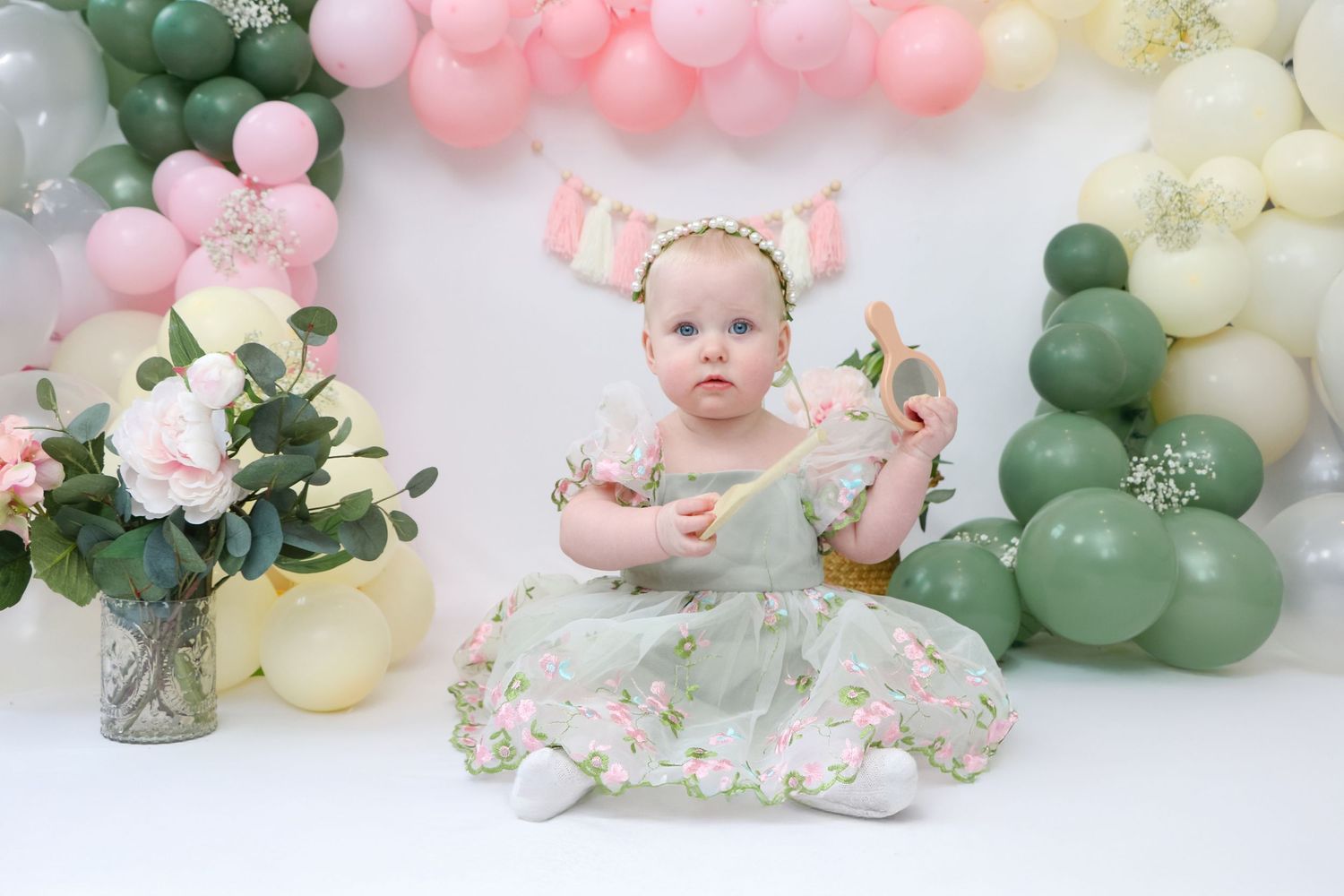 Baby girl in floral green dress sits before pink and green balloon arch with flowers for her first birthday cake smash.