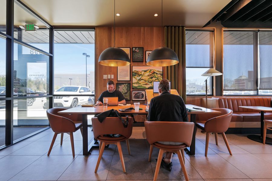 Two people sit at a wooden table in a modern restaurant with warm lighting, leather chairs, and large windows.