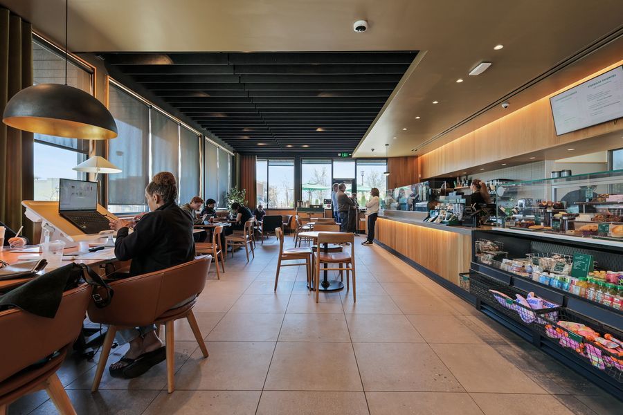 Modern Starbucks cafe interior with warm wood accents, pendant lights, customers seated, and a colorful display counter.