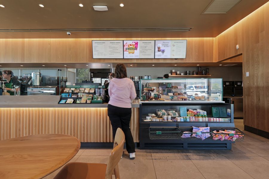 Woman ordering at a modern cafe counter with menu boards, display case of food items, and warm wood paneling.